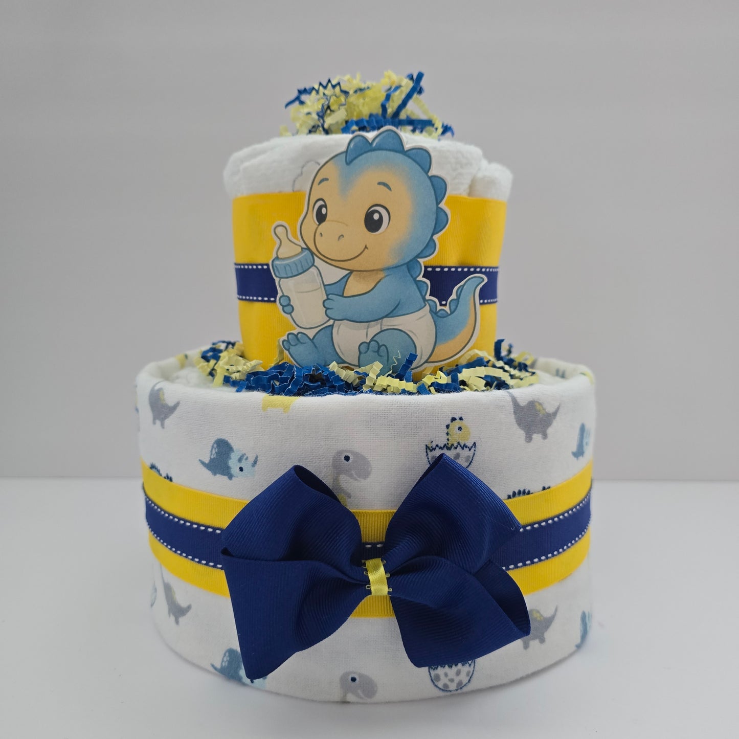 "Mini dinosaur diaper cake for baby boy with blue ribbon, small plush dinosaur, and stacked rolled diapers"