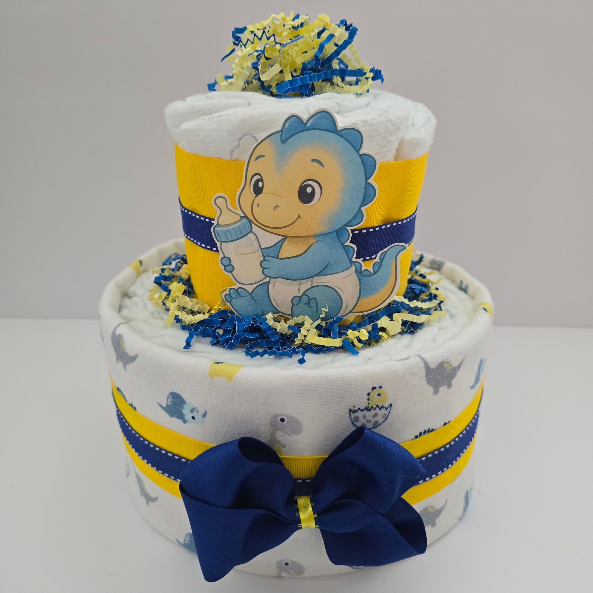 "Mini dinosaur diaper cake for baby boy with blue ribbon, small plush dinosaur, and stacked rolled diapers"